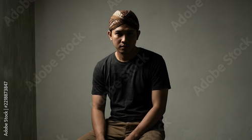 Confident young Indonesian man wearing a casual black t-shirt and traditional Javanese blangkon batik headpiece, blending modern lifestyle with cultural heritage in a dramatic studio portrait.