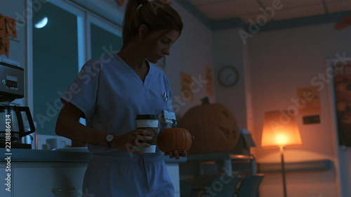 Young female nurse in blue medical scrubs holding a coffee cup and small pumpkin during a quiet halloween night shift break in a dimly lit hospital staff room
