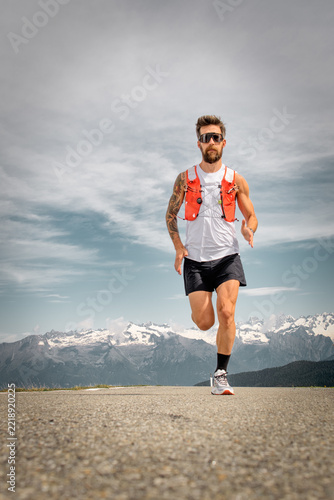 Athletic bearded man running on a mountain road wearing trail running gear against a scenic alpine landscape background