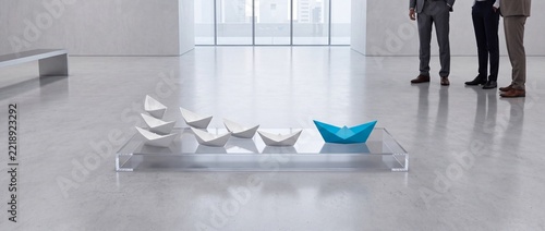 A single blue origami paper boat leading a row of white paper boats on a glass platform in a minimalist corporate gallery with businessmen standing in the background, symbol of leadership and