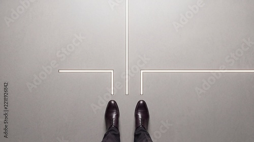 High-angle view of a professional businessman in leather shoes standing at a glowing pathway crossroad on a gray floor, symbolizing strategic decision-making, career growth, and premium success.