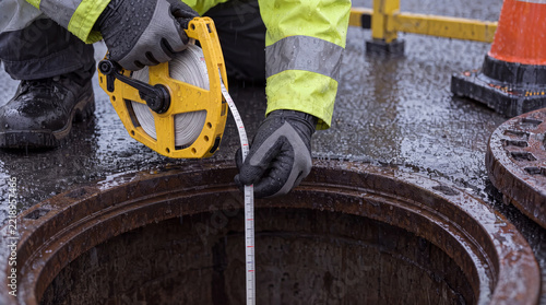 Utility Worker Measuring Manhole Depth with a Tape Measure in Wet Weather