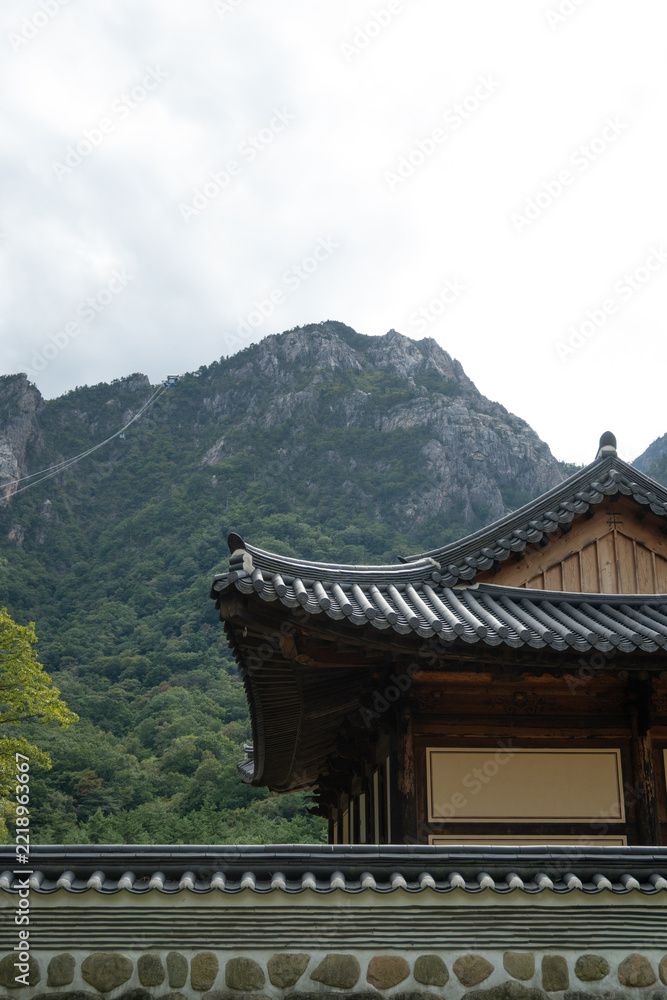 Obraz premium Traditional Korean temple roof in Seoraksan National Park with scenic rocky Seorak mountain peak and cable car line under overcast sky, historical Asian architecture and cultural travel background.