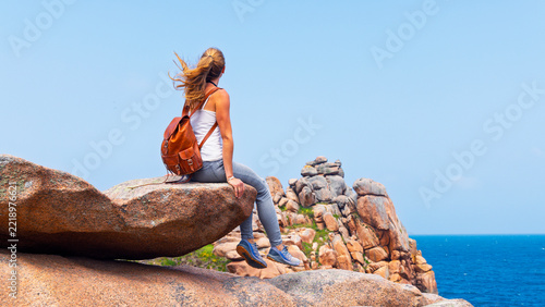 Young female hiker with leather backpack sitting on pink granite rock cliff overlooking ocean coast in Perros-Guirec Brittany France