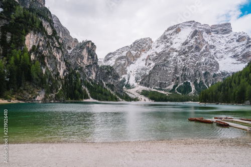 Turquoise alpine Lake Braies in the Dolomites, South Tyrol, Italy, with moored wooden rowboats, pebble shore, conifer forest and snow-capped limestone mountains under cloudy sky