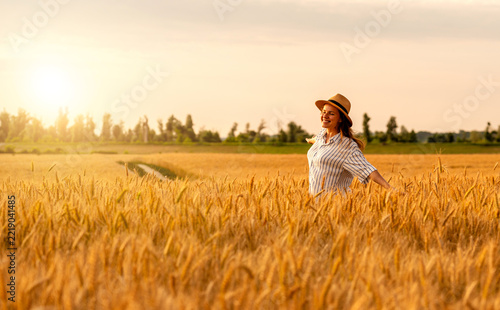 Cheerful woman wearing straw hat enjoying golden hour in wheat field in summer.