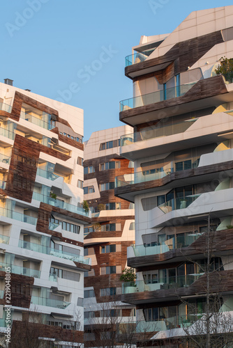 A low-angle view of contemporary apartment buildings frames a clear blue sky, highlighting layered balconies, angular façades, and the interplay of wood, glass, and pale stone