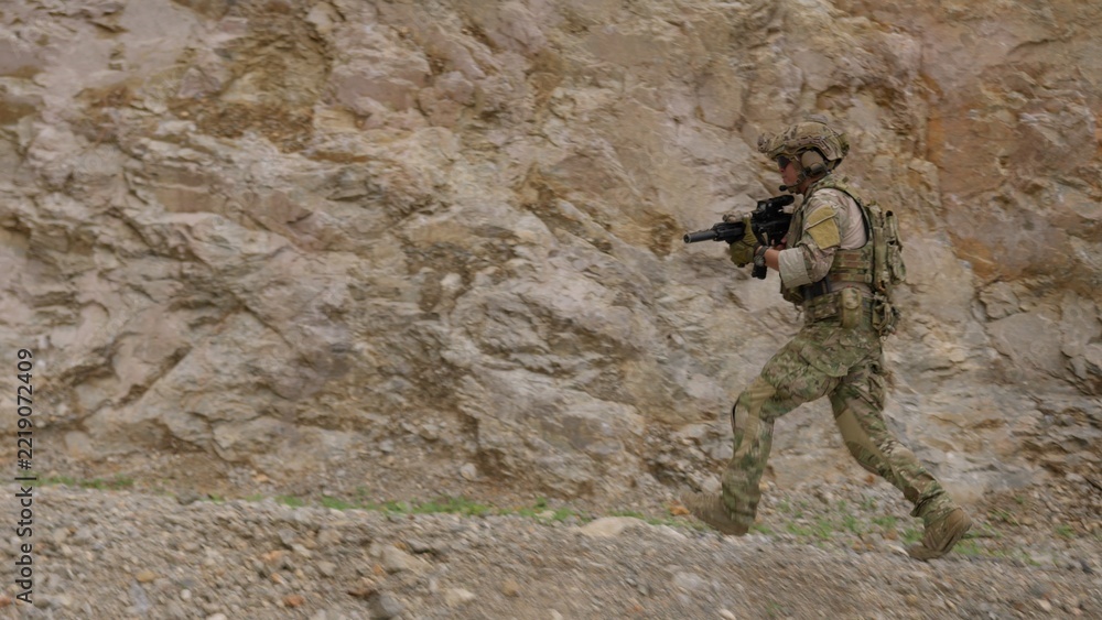 Fototapeta premium A special forces soldier in full combat gear advances with a rifle during a tactical mission, moving swiftly across rugged, rocky terrain with alertness and focus.