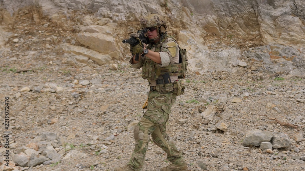 Fototapeta premium A special forces operator in full combat gear advances cautiously through a rocky, hostile environment while aiming his assault rifle on a tactical security patrol.