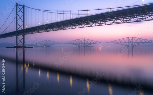 The Forth Road Bridge frames the historic Forth Bridge across a glassy Firth of Forth, under soft pink and violet pre-dawn light, with bridge lights reflected in the water.