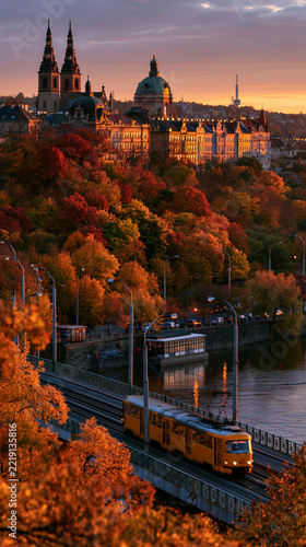 a cityscape during autumn. A yellow tram travels along a bridge over a river, its reflection shimmering on the waters surface. The city is bathed in warm hues of orange, red, and yellow.