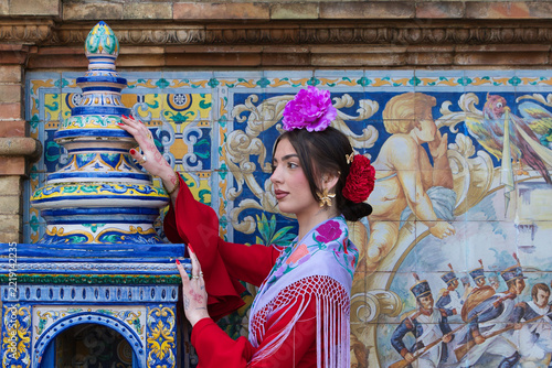 A beautiful flamenco dancer sitting on a tiled bench in a square in Seville, Spain. She is wearing a red ruffled dress and has a flower in her hair. Flamenco, a UNESCO Intangible Cultural Heritage.
