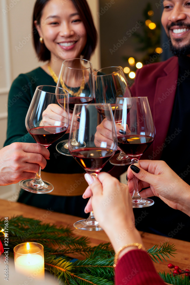 Fototapeta premium Vertical shot of diverse young adult friends toasting with red wine glasses during holiday celebration, useful for hospitality marketing and seasonal advertising campaigns