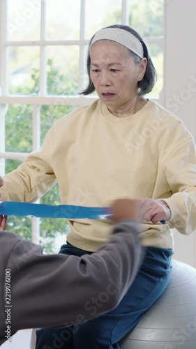 Vertical video: Pulling senior in yellow after PT holding blue band lifting arms rehab home on ball
