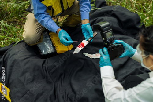 Obraz na plátně Forensic investigators in protective gloves photograph and examine a blood-stained knife on a black evidence cloth outdoors