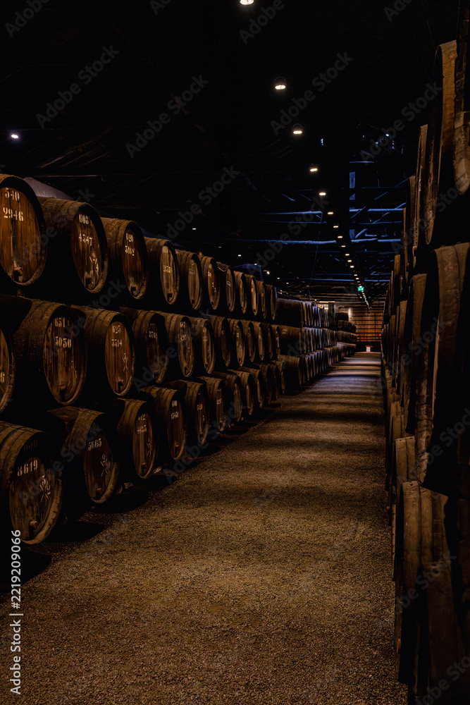Fototapeta premium Interior of traditional wine cellar in Porto Portugal with rows of wooden barrels used for aging port wine. Atmospheric underground storage with perspective view and warm low lighting.