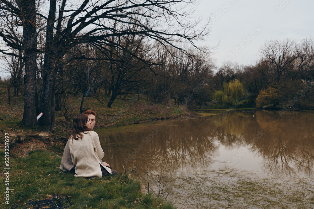 Naklejka premium Woman sits by a tranquil lake in a park wearing a cozy sweater, reflecting on a quiet day. Soft light and muted tones create a calm mood, highlighting nature and autumn ambiance.