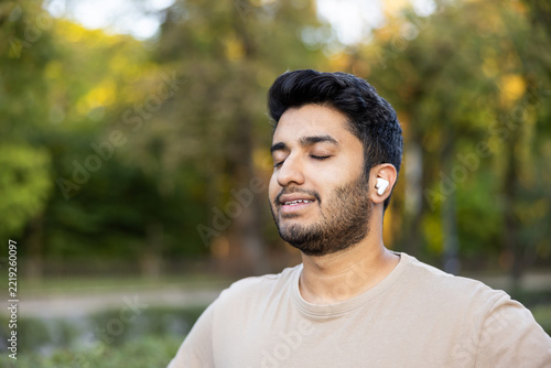 Young man with eyes closed, enjoying music or a podcast through wireless earbuds while relaxing in a sunlit park, peaceful, mindful, and content in nature