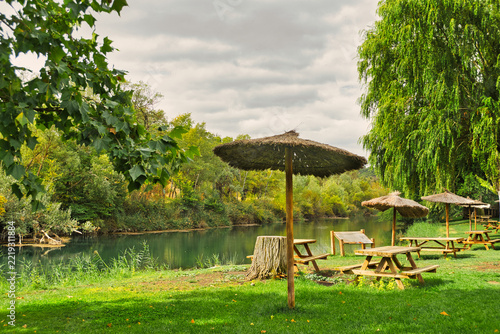 Fotografie Wooden picnic tables and thatched parasols beside the Tagus River, surrounded by willow trees in Zorita de los Canes, Guadalajara, Castilla-La Mancha, Spain