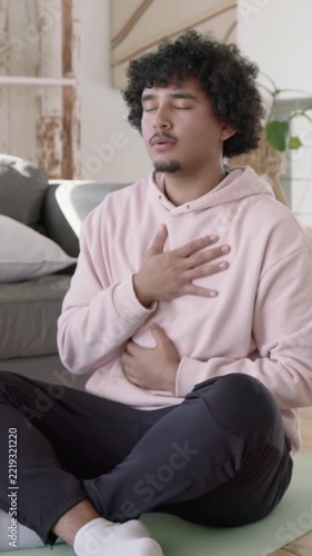 Vertical video: Man in pink hoodie placing hands chest and belly, breathing to relax on mat at home