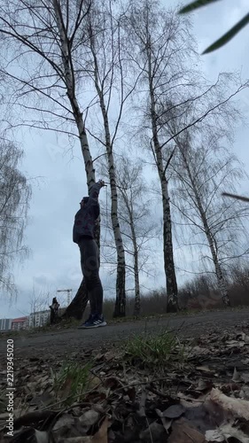 Man bending into deep warmup stretch under birch, hoodie and sneakers, leaf scattered ground, cloudy dull sky, vertical stock video showing prewalk routine and slow recovery