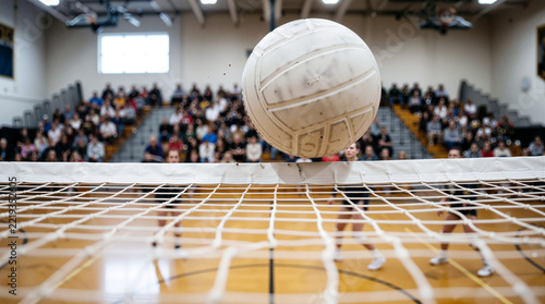Close-up of Volleyball Flying Over the Net During Indoor Match with Blurred Players and Spectators in Gymnasium