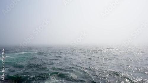 Dense sea fog rolling over dark, choppy ocean waters with sunlight glistening on the rolling waves.