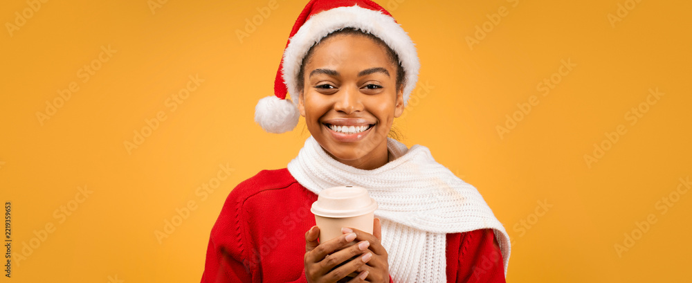 Fototapeta premium Happy Black Woman Smiling While Holding A Coffee Cup, Dressed In Festive Santa Hat And Scarf, Standing Against A Bright Yellow Background. Celebrating Holiday Spirit With Joyful Vibes.