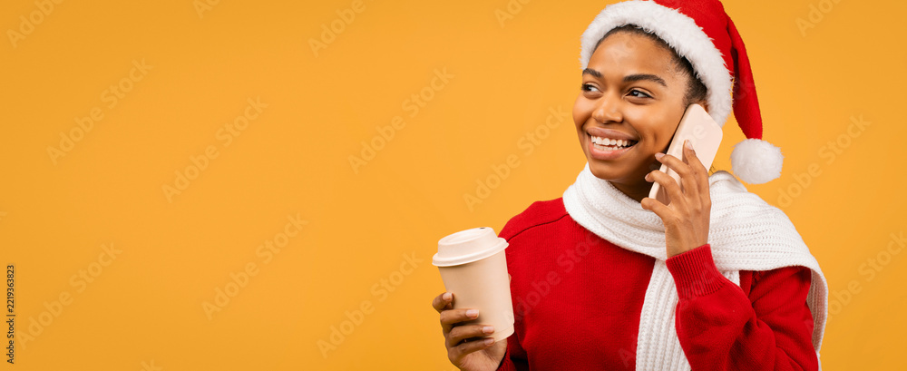Fototapeta premium Happy Black Woman In Santa Hat And Cozy Scarf Talking On Smartphone With Cup Of Coffee, Standing In Bright Indoor Setting. Joyful Mood Captured During Holiday Season, Celebration Concept.