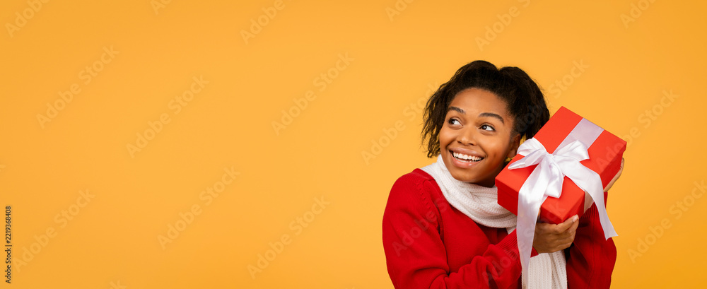 Fototapeta premium Smiling Young Black Woman Expressing Joy While Holding Red Gift Box With White Ribbon, Standing Against Bright Yellow Background, Signifying Celebration And Festivity, Gift Giving Concept.
