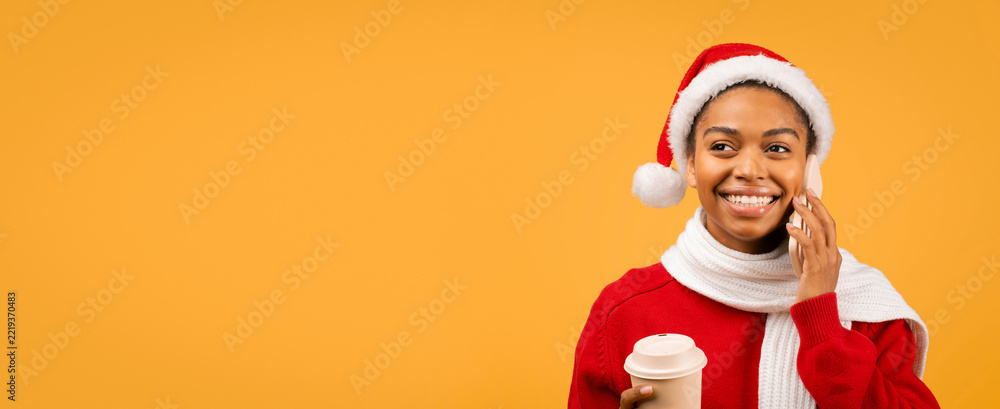 Fototapeta premium Smiling Young African American Woman Wearing Santa Hat While Talking On Phone With Coffee Cup In Hand, Standing Against Yellow Background, Embracing Holiday Spirit In A Festive Setting.