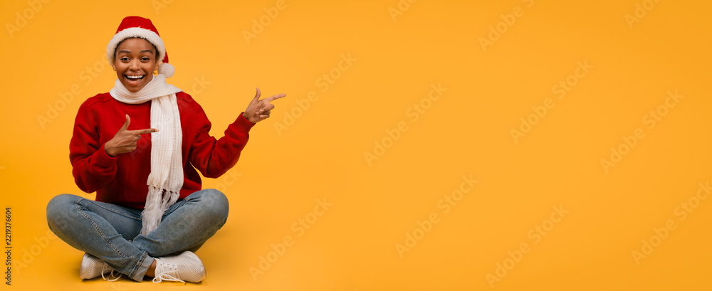 Fototapeta premium Smiling Black Woman Wearing Santa Hat Sitting Cross-legged On Studio Floor, Pointing Gesture With Hands In Festive Yellow Background, Expressing Cheerfulness And Joy For Christmas Celebration.