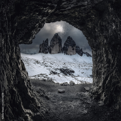 View of the Tre Cime di Lavaredo mountains framed by a dark rocky cave opening, with snow-covered slopes and a dramatic cloudy sky in Italy.