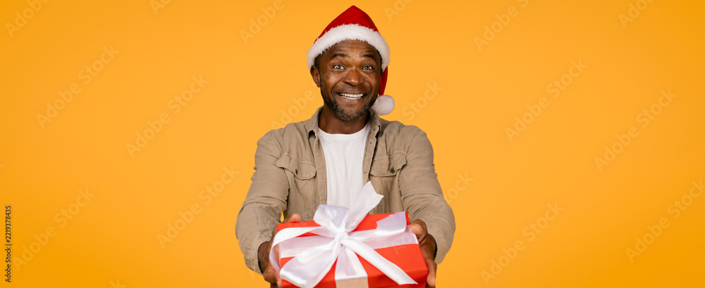 Fototapeta premium Smiling Black Male Adult In Santa Hat, Holding Red Gift Box With White Ribbon, Celebrating Christmas At Home. Joyful Emotion Indicates Happiness And Excitement For The Holiday Season.