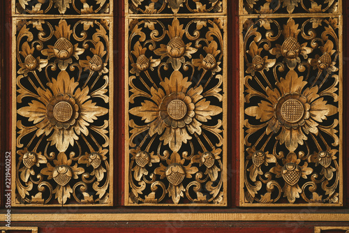 View of intricate traditional Balinese wood carvings with floral motifs and gold leaf detailing on a dark wooden panel in Ubud, Bali, Indonesia.