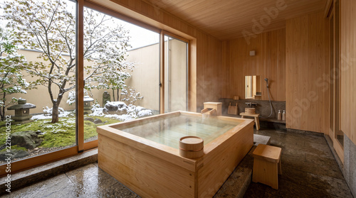 Luxury Japanese bathroom with steaming hinoki wooden bathtub and window view of snowy moss garden