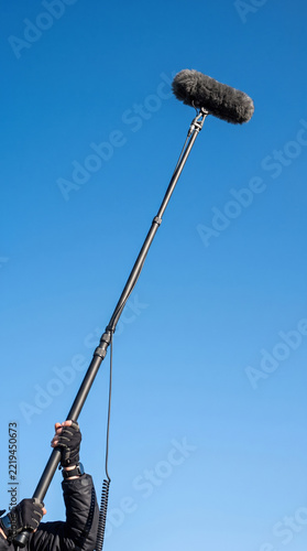 Long microphone reaching into blue sky above person holding boom pole outdoors