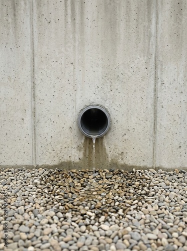 Water Draining from Plastic Pipe into Gravel Bed against Raw Concrete Wall, Industrial Drainage System Concept, Minimalist Architectural Detail, Urban Infrastructure Maintenance, Environmental Drainag