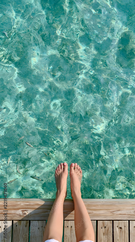 Fototapeta premium Bare legs dangling over clear turquoise water beside wooden pier, creating relaxed summer mood in tropical swimming spot