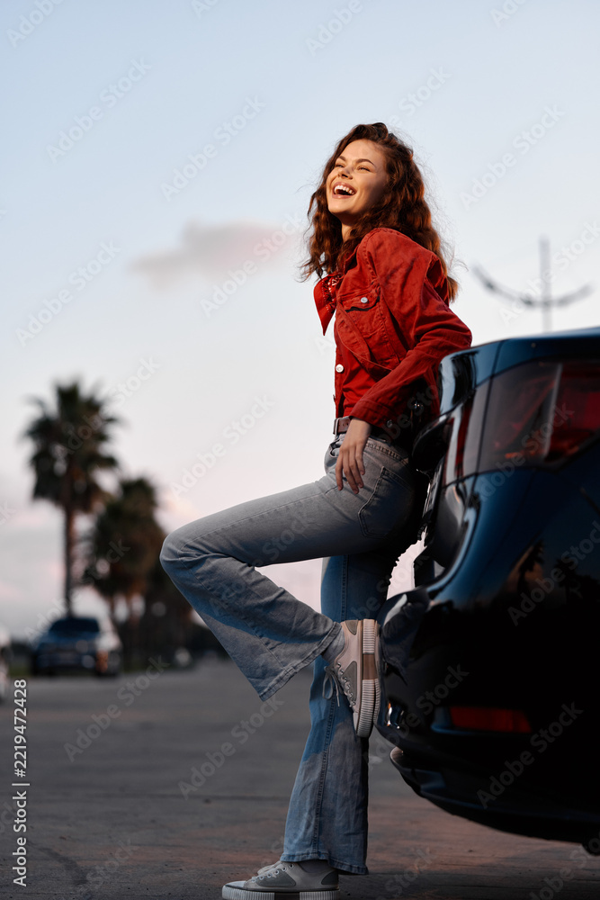 Fototapeta premium Smiling woman leaning against car. Young laughing female outdoors. Concept of freedom, travel, and road trip. Sunset lighting creating warm mood.
