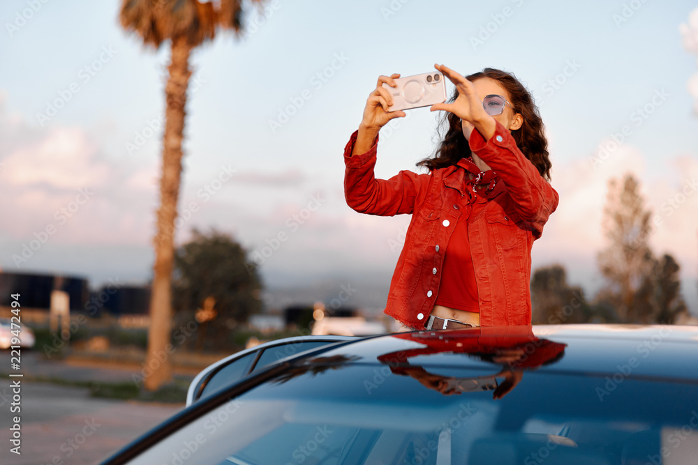 Naklejka premium Young woman taking photo with smartphone standing through car sunroof. Sunset light, travel and modern lifestyle concept, palm tree background.