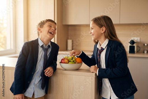 Happy school children laughing in kitchen before school, pupils wearing uniform eating fresh fruit snack, siblings morning routine
