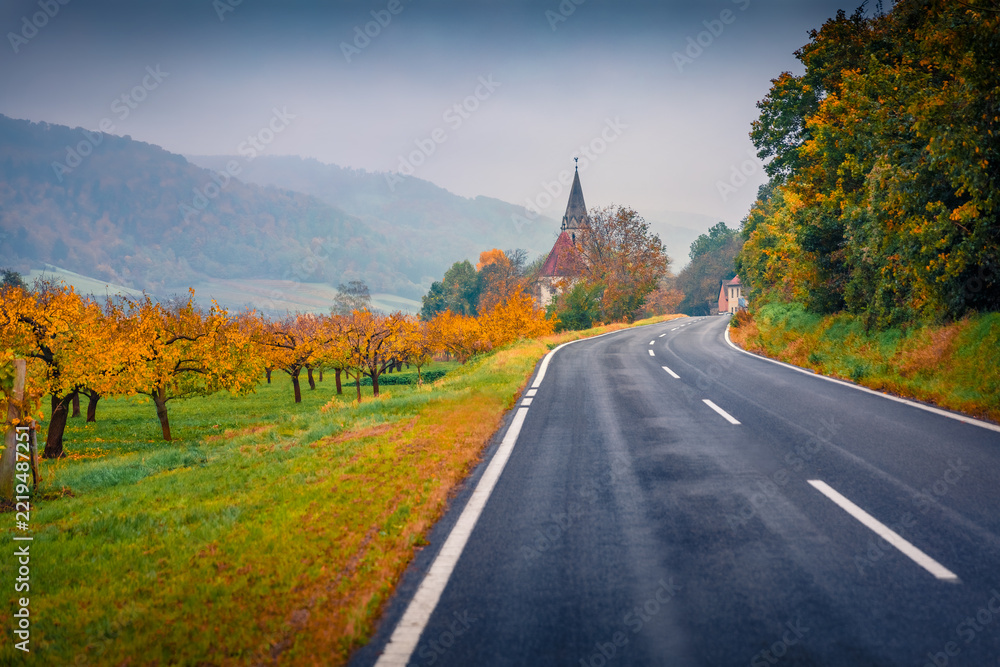 Fototapeta premium Spectacular autumn view of apple tree garden in a mountain valley with St. Johann in Mauerthale Catholic church on background, Austria. Rainy morning scene of empty asphalt road in the austrian Alps.