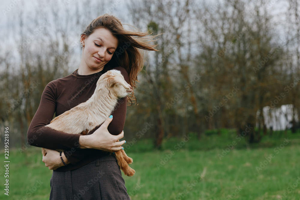 Naklejka premium A woman in a brown long sleeve top gently hugs a goat kid in an open field, wind in her hair, smiling softly. A warm, candid lifestyle moment of bonding, care, and rural nature.