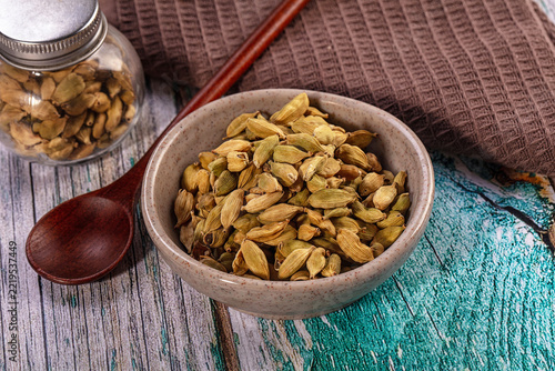 Wooden spoon beside ceramic bowl filled with green cardamom pods on weathered blue and gray wooden surface, spice ingredient