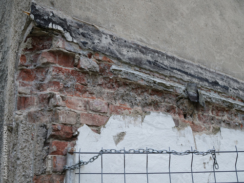 damaged exterior building wall where the top layers of grey stucco and white plaster have broken away, exposing a crumbling red brick foundation and a degraded horizontal bitumen barrier behind a wire