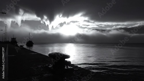 Dramatic black and white seascape with sun bursting through clouds above calm water and boats at the harbor