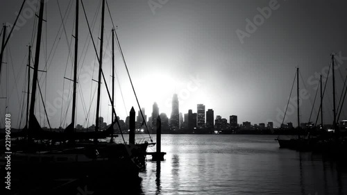 Black and white view of sailboats in a harbor silhouetted by the cityscape at dusk or dawn.