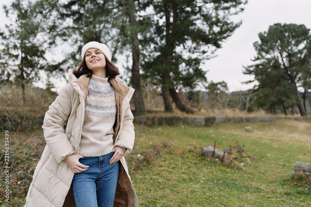 Naklejka premium Woman in a cozy coat and knit sweater posing outdoors in a park, with trees and open fields behind her.