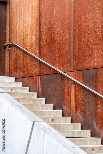 Concrete stairs with a diagonal metal handrail alongside a rust colored steel wall showing material contrast in modern exterior architecture